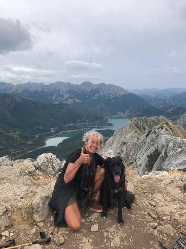 A joyful woman kneels beside her black dog on a rocky mountaintop, with dramatic mountains, a winding river, and cloudy sky creating a breathtaking scenic backdrop.
