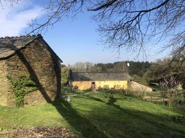 A rustic stone building and a yellow cottage sit in a sunny, grassy yard surrounded by trees and fields, under a bright blue sky.