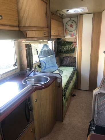 This image shows the interior of a cozy camper van, featuring a compact kitchen, wooden cabinets, bed with pillows, dreamcatcher, and a small circular sink.