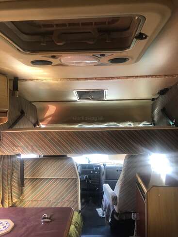 This image shows the interior of a camper van, featuring striped upholstery, a compact dining area, overhead bunk bed, front driving seats, and natural sunlight entering.