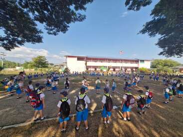 Cette image montre un grand groupe d'enfants en uniforme scolaire se tenant à l'extérieur sur un terrain, espacés les uns des autres, avec un bâtiment scolaire et un drapeau à l'arrière-plan.