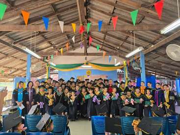 Un grand groupe de diplômés, vêtus de toges et de casquettes noires, posent pour une photo de célébration à l'intérieur, tenant des certificats et des animaux en peluche, sous des drapeaux colorés suspendus.