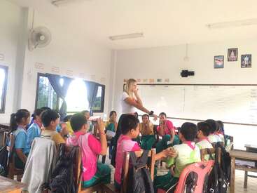 Un enseignant se tient à l'avant d'une salle de classe bien éclairée et discute avec un groupe d'élèves attentifs, assis en cercle, qui participent avec enthousiasme.
