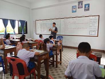 Un professeur se tient à l'avant d'une salle de classe, un livre à la main, tandis que de jeunes élèves en uniforme blanc sont assis à des pupitres et écoutent et écrivent attentivement.