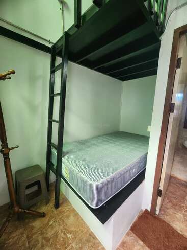 Une petite chambre à coucher confortable avec un matelas sur une plate-forme surélevée, une échelle noire, un porte-manteau en bois, un tabouret, un sol carrelé et un décor minimaliste sous un escalier.