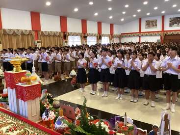 Un grand groupe d'étudiants en uniforme se tient dans un hall décoré, les mains jointes en signe de respect, participant à une cérémonie formelle avec des offrandes à l'avant.