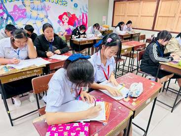 Dans une salle de classe à la décoration lumineuse, des élèves en uniforme travaillent assidûment à leur bureau, rédigeant et collaborant à des travaux, créant ainsi une atmosphère de concentration et d'apprentissage en commun.