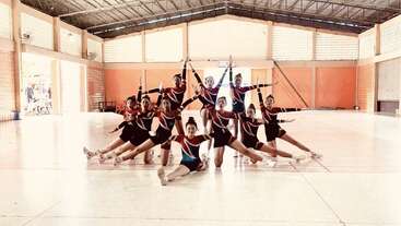 Une équipe de pom-pom girls pose dans un grand gymnase couvert, portant des uniformes assortis. Elles forment une formation synchronisée, souriant avec assurance, bras et jambes tendus à l'unisson.