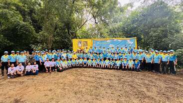Un grand groupe de personnes vêtues d'uniformes bleus et d'écharpes jaunes pose ensemble devant une bannière jaune et bleue, entourée d'arbres verdoyants en plein air.