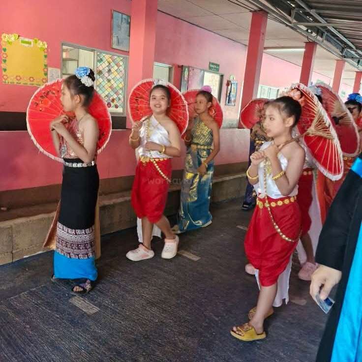 A group of young girls dressed in traditional Thai clothing walk along a corridor, holding red parasols. They appear to be participating in a cultural event or celebration.