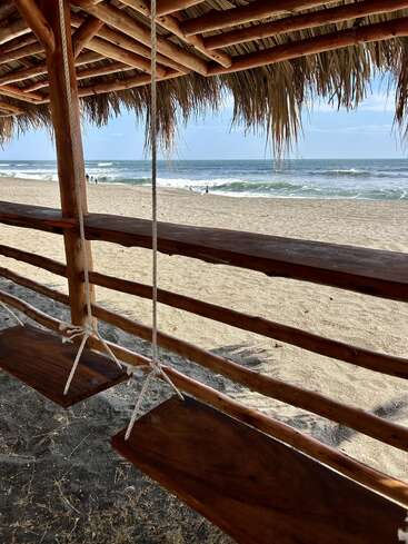 Under a thatched-roof hut, two wooden swings hang facing a sandy beach and ocean waves. People walk near the water under a bright, serene sky.