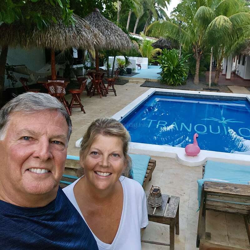 Una pareja sonriente posa para una selfie junto a una piscina. Las palmeras tropicales, las cabañas con techo de paja y los muebles de madera crean un ambiente relajante, similar al de un resort vacacional.