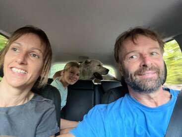 Uma família sorridente está sentada dentro de um carro: uma mulher, um homem, uma menina e um cachorro feliz no banco de trás, aproveitando a viagem juntos.