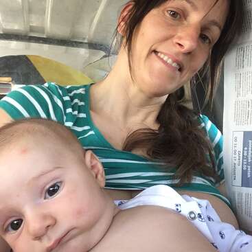 A woman in a green striped shirt smiles while holding a baby close to her chest. The baby looks calm. A newspaper is partially visible.