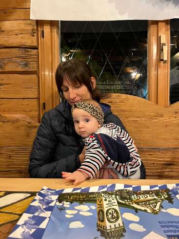 Uma mulher segura carinhosamente um bebê usando uma faixa na cabeça e uma roupa listrada. Eles estão sentados em um restaurante aconchegante de madeira, com jogos americanos coloridos e iluminação aconchegante ao redor.