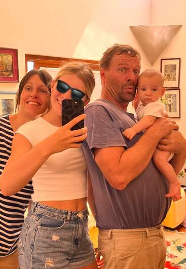 Uma família feliz tira uma selfie no espelho. A garota da frente usa óculos escuros, sorrindo, enquanto o pai segura um bebê. Atmosfera calorosa, aconchegante e alegre.