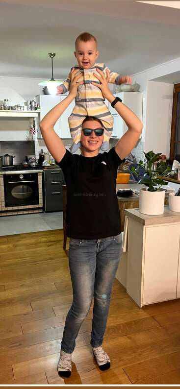 Um adulto sorridente usando óculos escuros segura um bebê feliz nos ombros em uma cozinha aconchegante. Ambos parecem alegres, compartilhando um momento lúdico e amoroso juntos.