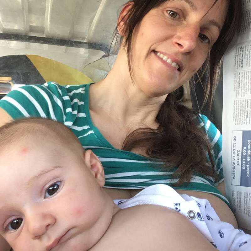Uma mulher com uma camisa listrada verde sorri enquanto segura um bebê junto ao peito. O bebê parece calmo. Um jornal está parcialmente visível.