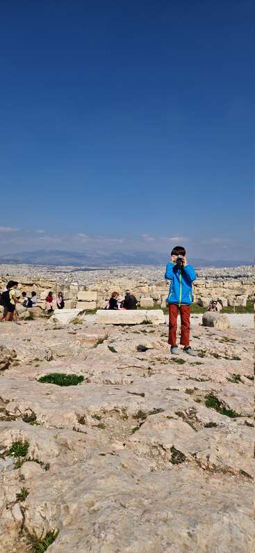 Un enfant vêtu d'une veste bleue se tient sur d'anciennes ruines en pierre et photographie le paysage. Plusieurs personnes s'assoient et marchent à proximité, avec une vaste ville et des montagnes au-delà.