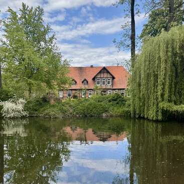 The image depicts a serene scene of a large house with a red roof, situated behind a tranquil pond, surrounded by lush trees and greenery on a sunny day.