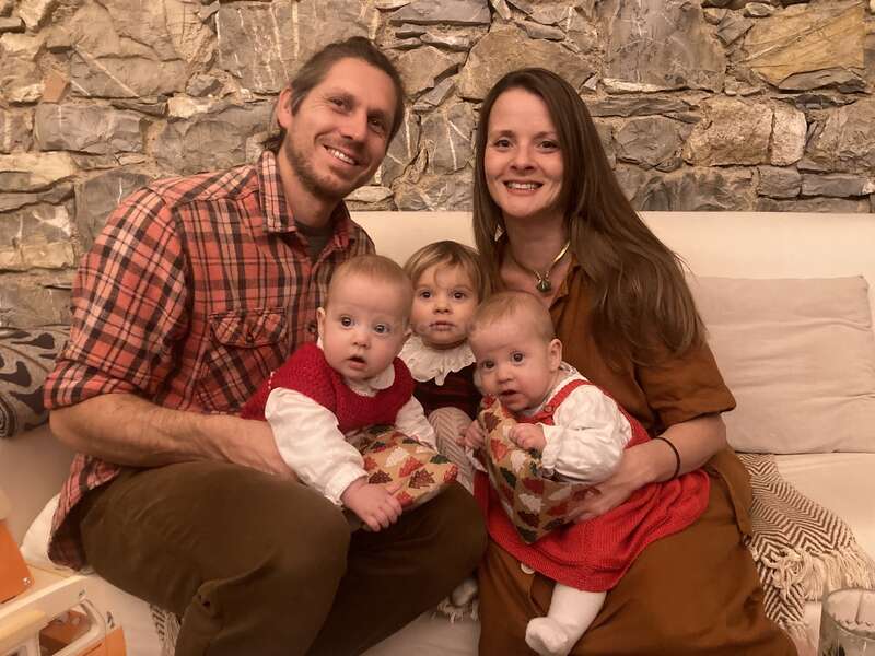 A smiling family of five sits on a couch in front of a stone wall. The parents hold two babies, while a toddler sits between them.