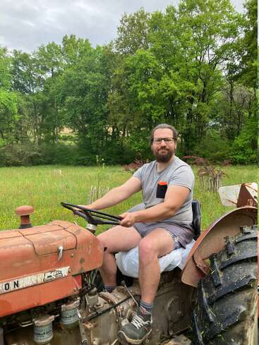 Un hombre barbudo con gafas, pantalones cortos y camiseta está sentado en un viejo tractor en un frondoso campo verde con árboles bajo un cielo nublado.