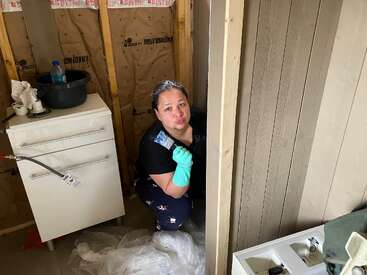 Una mujer con guantes y pijama está pintando o renovando una habitación. Posa juguetonamente, rodeada de materiales de construcción, aislantes y herramientas, creando una atmósfera informal y trabajadora.