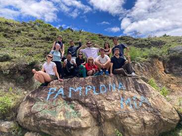 Um grupo de doze amigos posa em uma encosta ensolarada atrás de uma grande rocha pintada com "Pamplona Mía", cercada por vegetação e céu azul com nuvens.