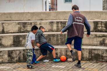 Três meninos estão jogando futebol em uma área pavimentada com degraus de concreto como fundo. Duas crianças desafiam um menino mais velho pela bola laranja, desfrutando de um jogo enérgico.