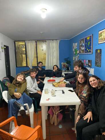 Um grupo de amigos está reunido em torno de uma mesa branca, sorrindo e posando para uma foto. Uma pessoa se esconde de brincadeira embaixo da mesa, em uma sala bem iluminada.