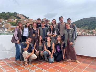 Um grupo de dezesseis jovens adultos posa alegremente em um telhado com montanhas e uma cidade ao fundo. A maioria usa coletes semelhantes, sugerindo trabalho em equipe ou organização.
