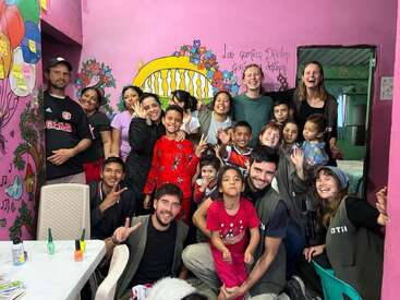 Um grupo alegre de adultos e crianças posa junto em uma sala colorida com paredes cor-de-rosa, obras de arte animadas e rostos sorridentes, irradiando felicidade e união.