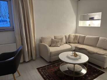 This cozy living room features a cream sectional sofa, round coffee table, armchair, decorative rug, warm lighting, a window with curtains, and minimalist wall shelving.