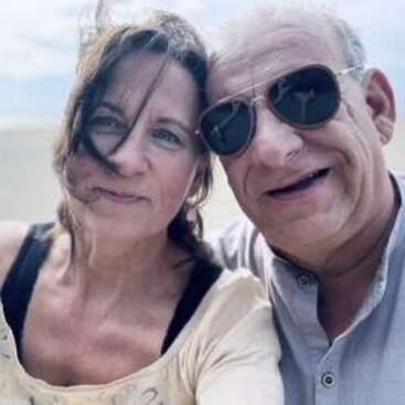 Ein Paar mittleren Alters macht draußen ein Selfie aus der Nähe. Die Frau hat vom Wind zerzaustes Haar, während der Mann eine Sonnenbrille trägt und lächelt. Der Hintergrund sieht hell und sandig aus.