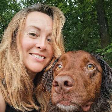 A woman with long, light brown hair smiles while taking a selfie with her brown dog. They are outdoors, surrounded by lush, green trees in the background.