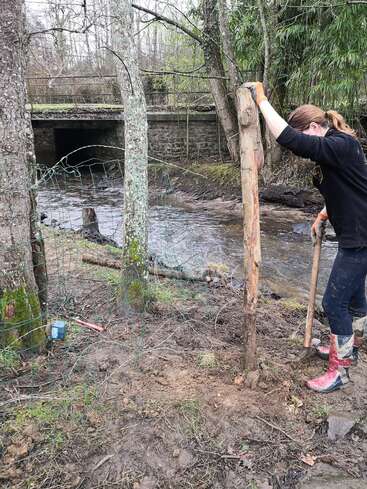 Uma pessoa com botas e luvas vermelhas cava perto de um rio, consertando ou instalando uma cerca de arame perto de árvores, com uma ponte de pedra visível ao fundo.