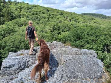 Um homem com uma mochila está em um afloramento rochoso ao lado de um cachorro marrom, com vista para uma floresta verde e exuberante sob um céu parcialmente nublado, apreciando a natureza.