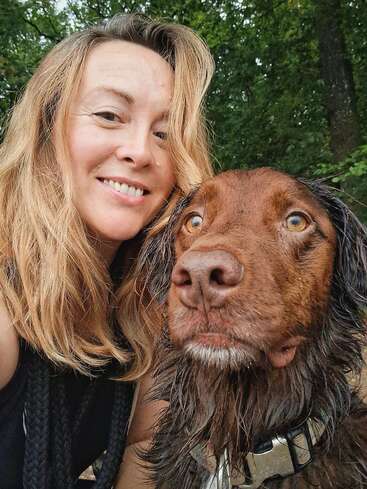 Uma mulher com longos cabelos castanhos claros sorri enquanto tira uma selfie com seu cachorro marrom. Eles estão ao ar livre, cercados por árvores verdes e exuberantes ao fundo.