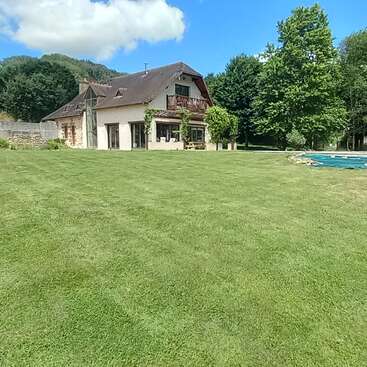Ein hübsches Haus auf dem Land mit einem Schrägdach, umgeben von einem großen, gepflegten grünen Rasen, Bäumen, blauem Himmel und einem kleinen Swimmingpool.