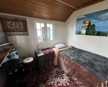 Ein gemütliches Schlafzimmer im Dachgeschoss mit einem Einzelbett, einem gemusterten Teppich, Musikinstrumenten, einem Kunstwerk mit einer goldenen Kuppel, einem Fenster, einer Holzdecke und einem dekorativen Wandbehang.