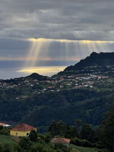 Goldene Sonnenstrahlen durchdringen dramatische Wolken und beleuchten ein friedliches Küstendorf inmitten üppig grüner Hügel, während das Meer das Licht unter einem ruhigen Himmel schimmert.