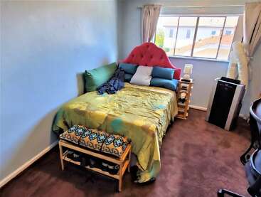 A cozy bedroom with light blue walls, a bed with colorful pillows, pink headboard, side tables, a window, bench, and an air conditioning unit.