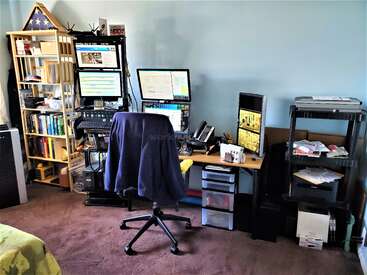 Das Bild zeigt eine Homeoffice-Einrichtung mit mehreren Monitoren, einem Schreibtischstuhl, Bücherregalen, Büromaterial, verschiedenen elektronischen Geräten, Büchern und übersichtlichen Aufbewahrungsmöbeln vor einer blauen Wand.