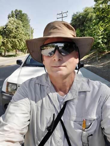 Ein Mann mit einem breitkrempigen Hut, einer Sonnenbrille und einem hellen Hemd macht draußen ein Selfie, während er an einem sonnigen Tag vor einem weißen Auto steht.