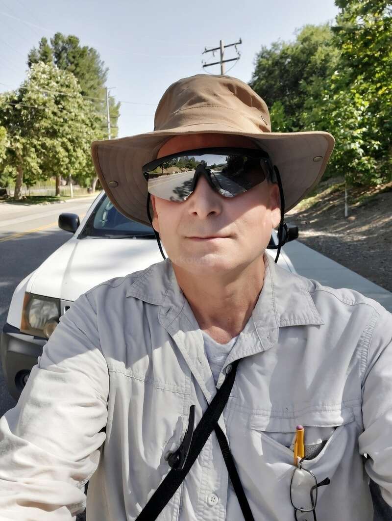 Ein Mann mit einem breitkrempigen Hut, einer Sonnenbrille und einem hellen Hemd macht draußen ein Selfie, während er an einem sonnigen Tag vor einem weißen Auto steht.