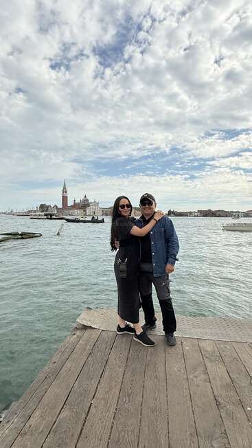 Un couple pose sur un quai en bois au bord de l'eau, avec en arrière-plan l'architecture vénitienne historique et des bateaux, sous un ciel nuageux spectaculaire. Paisible et pittoresque.