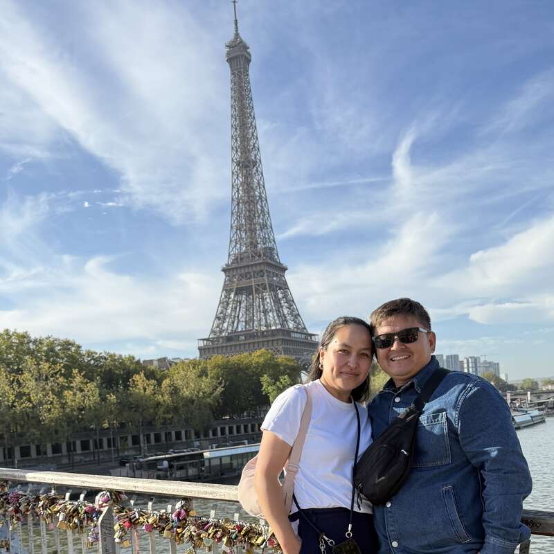 Un couple souriant pose sur un pont à Paris, avec la tour Eiffel et un magnifique ciel bleu en arrière-plan, entouré de cadenas d'amour.