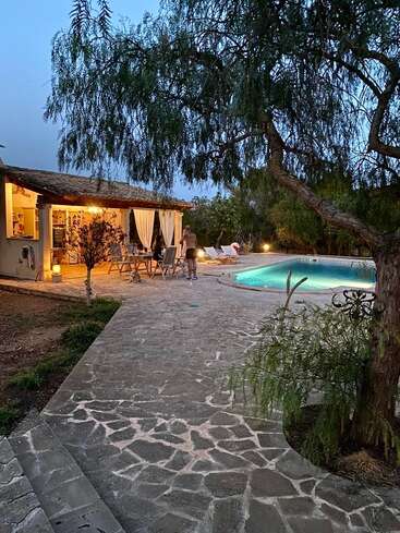 Das Bild zeigt eine gemütliche Terrasse mit einem Swimmingpool, umgeben von Bäumen. Die Menschen entspannen sich unter warmen Lichtern, während der Abend hereinbricht und eine friedliche Atmosphäre schafft.