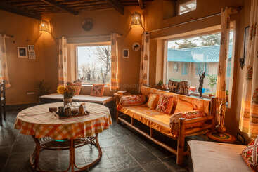 This cozy living room features large windows, wooden furniture, warm lighting, patterned cushions, yellow tones, indoor plants, and a round table with a checkered cloth.