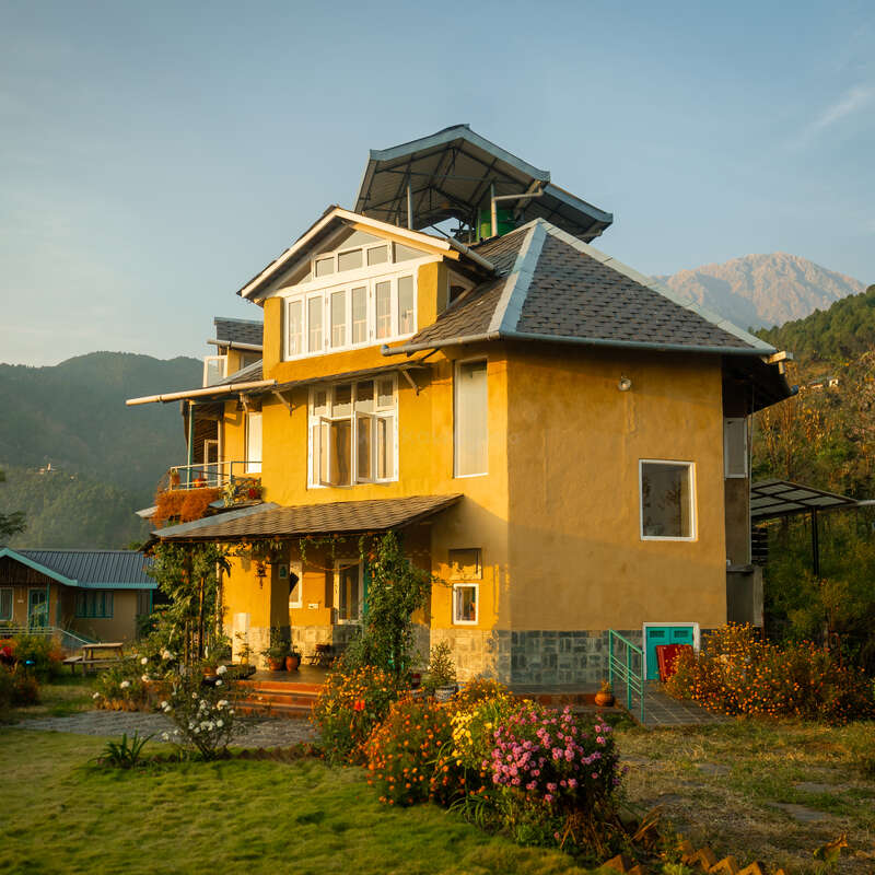 A charming yellow house with large windows, surrounded by colorful flowers and greenery, set against scenic mountains under a clear sky, bathed in warm sunlight.